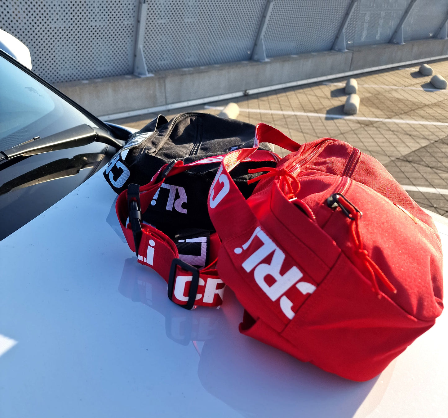 Sac bandoulière rouge CRLi posé près d’une voiture, logo et sangle