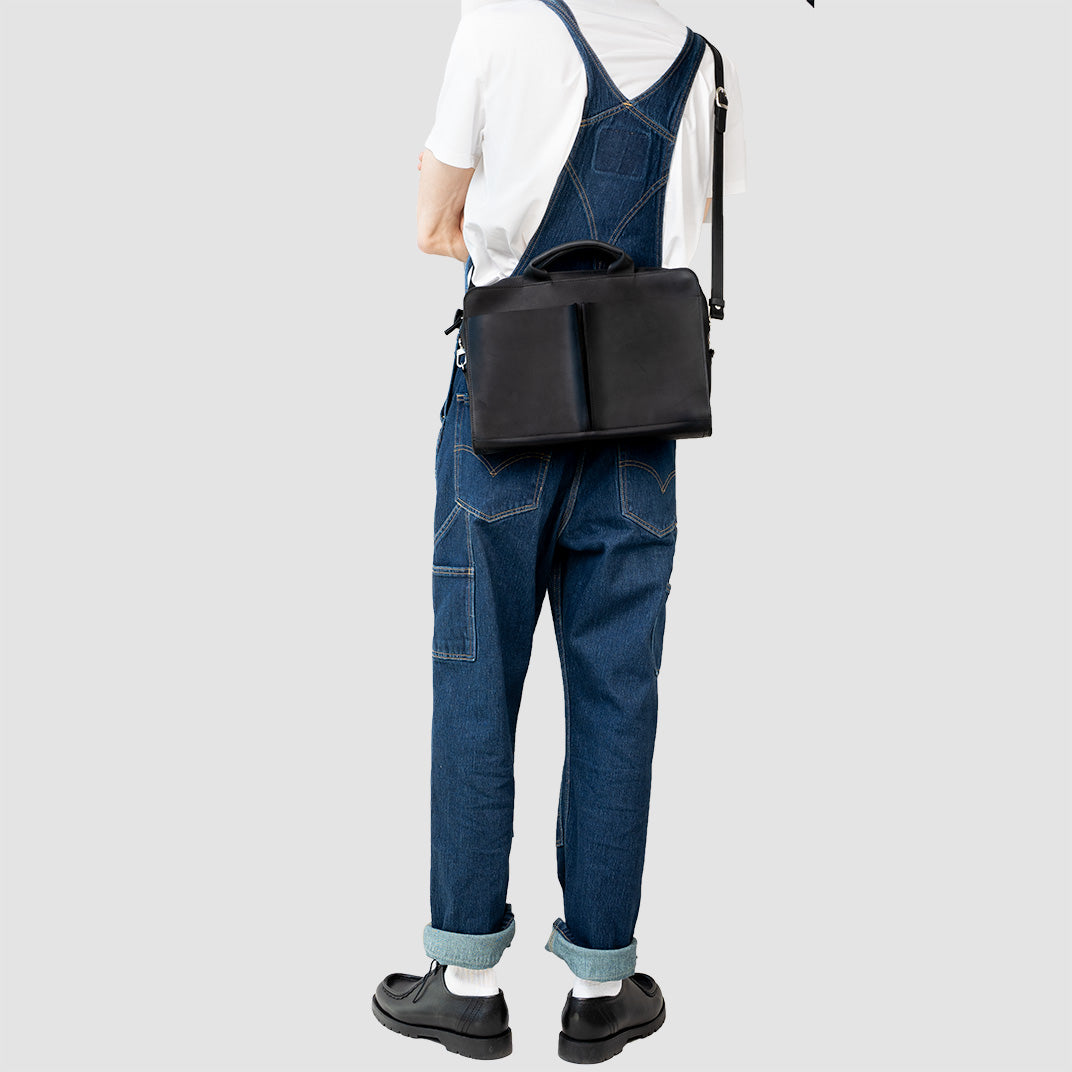 Sac ordinateur en cuir noir en bandoulière, porté sur jean et t-shirt blanc