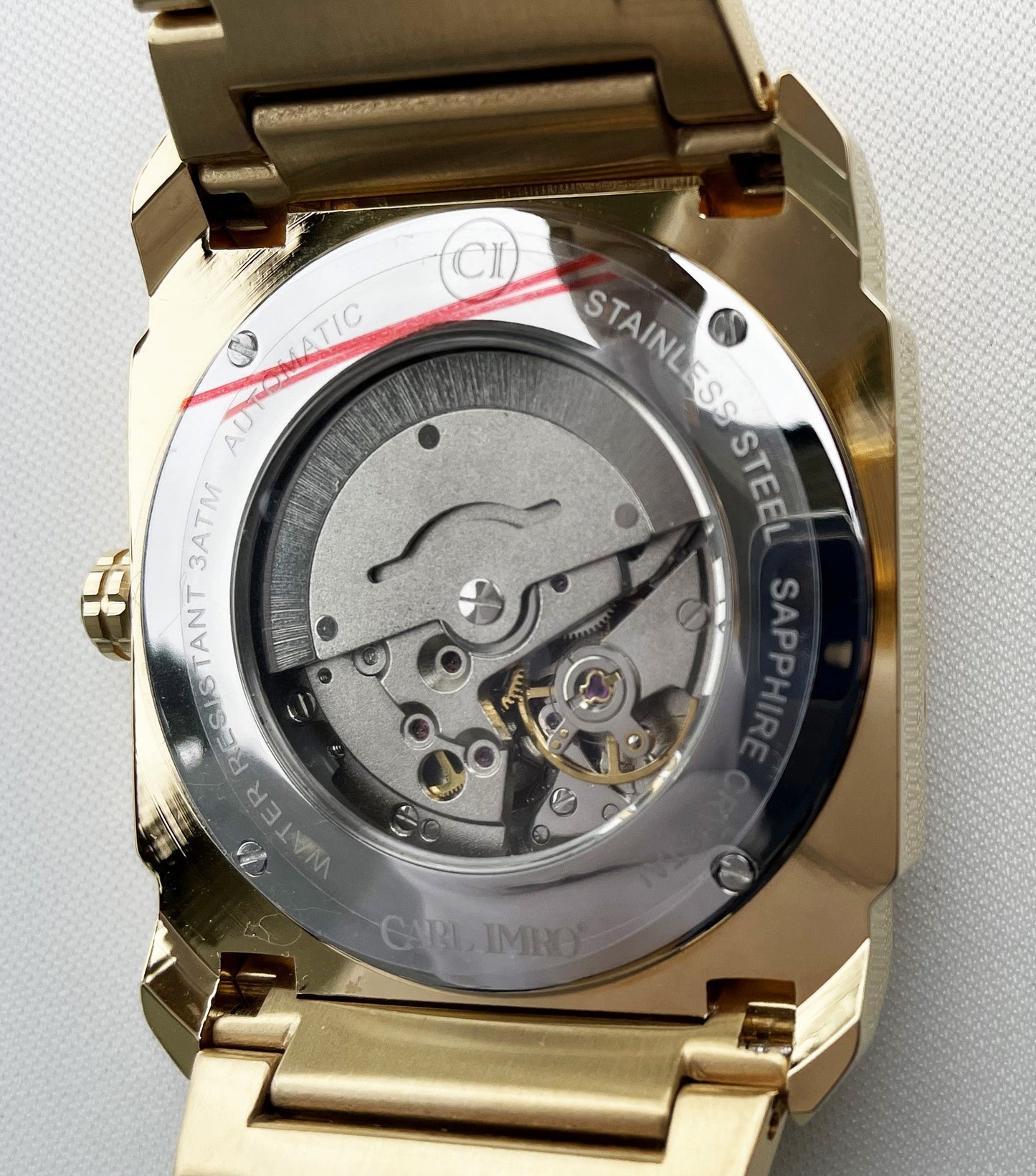 Montre-bracelet acier doré ouverte, mécanisme gris visible, marquages sur le fond.