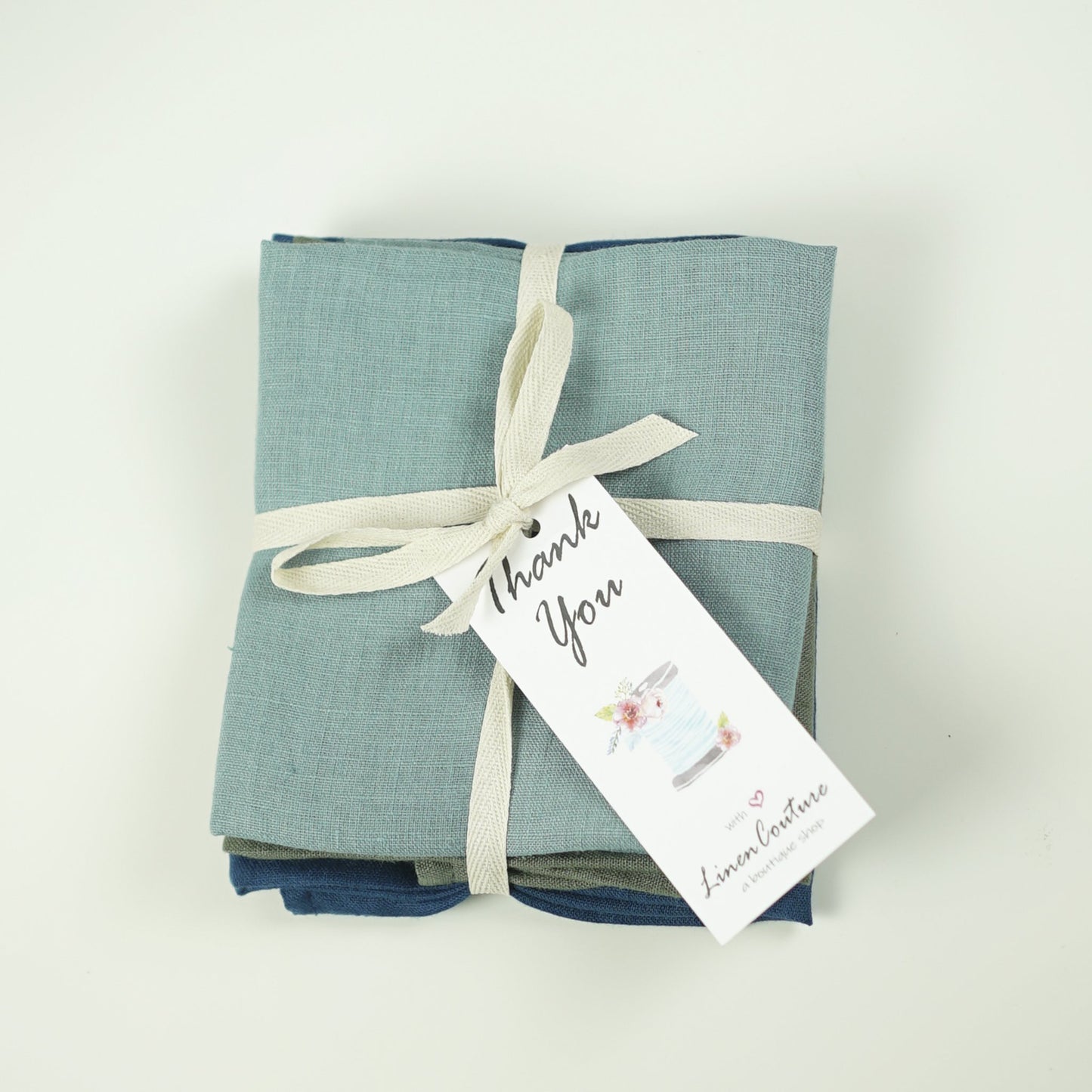 Sac fourre-tout en lin bleu-vert plié, ruban crème et étiquette cadeau.