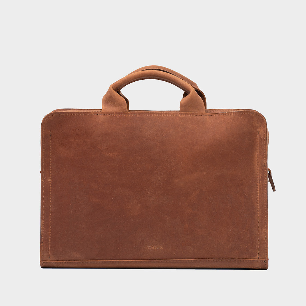 Sac d’ordinateur en cuir cognac à anses, surface lisse, zip sur le côté.